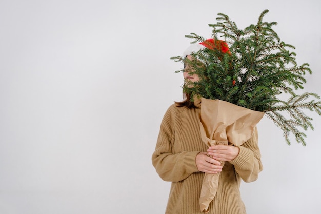 Una giovane donna caucasica sorridente che indossa il cappello di Babbo Natale che fa capolino da dietro i rami dell'albero di Natale guardando la telecamera su sfondo bianco isolato