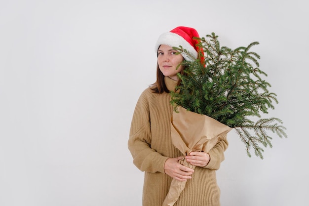 Una giovane donna caucasica sorridente che indossa il cappello di Babbo Natale che fa capolino da dietro i rami dell'albero di Natale guardando la telecamera su sfondo bianco isolato