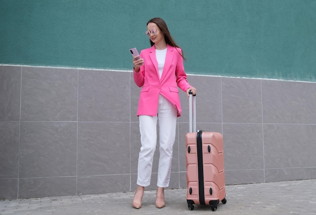 Una giovane donna che indossa una giacca rosa brillante sta con una valigia e un telefono nelle sue mani concetto di viaggiatore