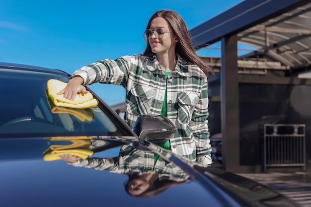 Una giovane donna che pulisce e lucida la sua auto con un panno in microfibra giallo Concetto di cura dell'auto