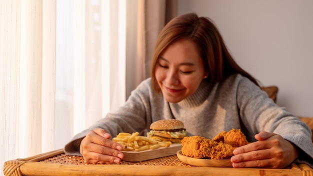 Una giovane donna che tiene e mangia un hamburger patatine fritte e pollo fritto sul tavolo a casa