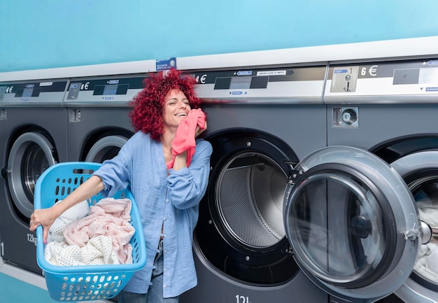 Una giovane donna con capelli afro rossi che odorano di vestiti puliti che tengono un cestino con i vestiti in una lavanderia automatica blu