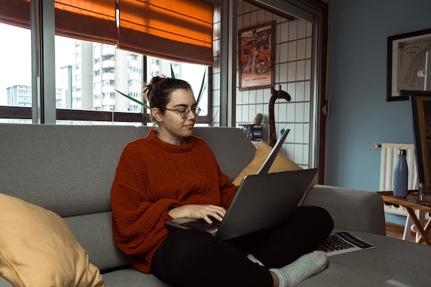 Una giovane donna con gli occhiali che lavora e studia sul suo laptop sul divano di una casa moderna