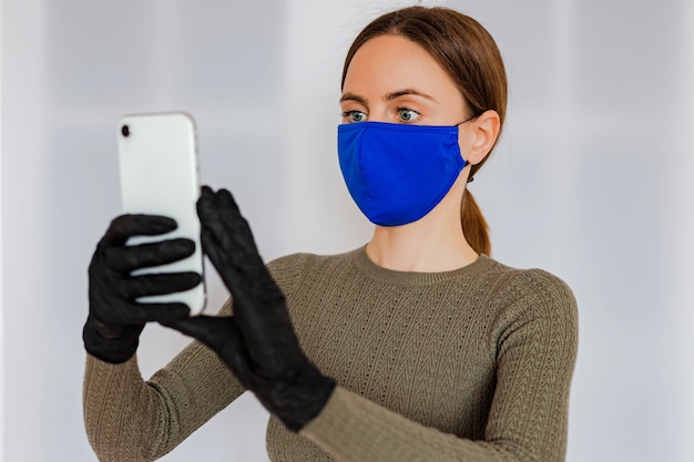 Una giovane donna con i capelli castani chiari con uno smartphone bianco in mano in guanti neri di lattice medico, che indossa una maschera medica di cotone blu