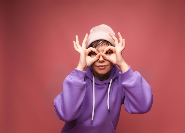 Una giovane donna con i capelli corti in un cappello mostra gli occhiali con le mani guarda direttamente nella fotocamera alla ricerca di qualcosa Ritratto di una ragazza divertente in jeans su sfondo rosa