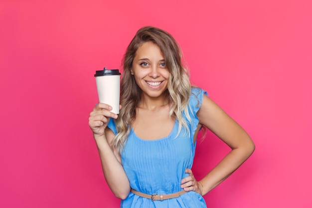 Una giovane donna con i capelli ondulati tiene in mano una tazza di caffè o tè di carta bianca su uno sfondo rosa