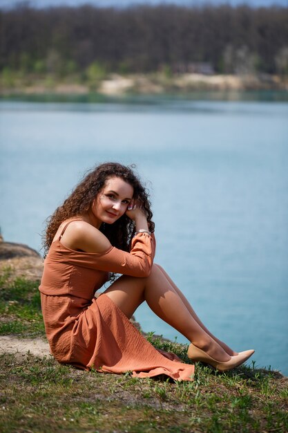 Una giovane donna con i capelli ricci e un sorriso sul viso siede in un prato verde sullo sfondo di un lago blu. Calda giornata estiva, ragazza felice, emozioni di gioia