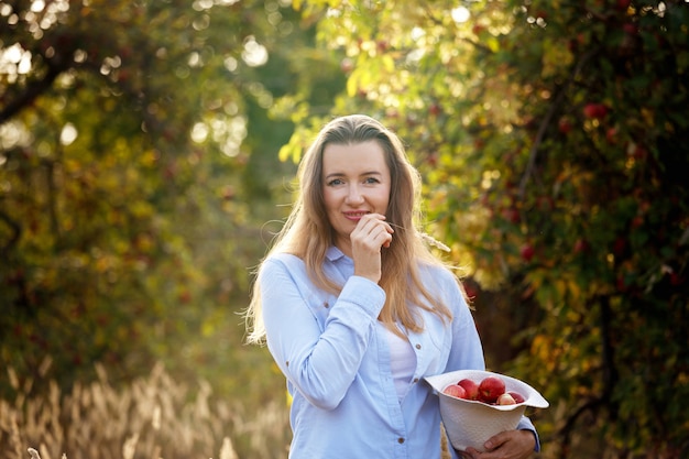 Una giovane donna con le mele in un cappello è in piedi in un giardino delle mele in una giornata di sole autunnale. Concetto di stile di vita sano