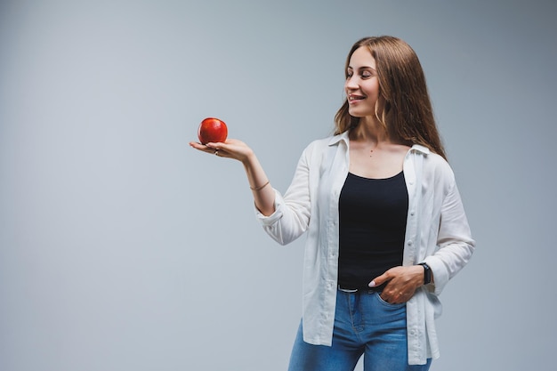 Una giovane donna con lunghi capelli castani, vestita con una camicia bianca e jeans, tiene in mano una mela rossa fresca. Concetto di cibo sano Sfondo bianco