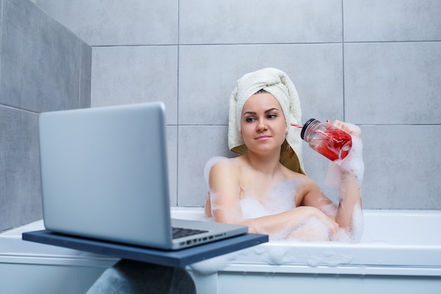 Una giovane donna con un asciugamano in testa beve succo di frutta guardando un film su un laptop mentre è seduta in un bagno in un salone di bellezza. Rilassati in bagno senza biancheria. Cura del corpo e concetto di relax.