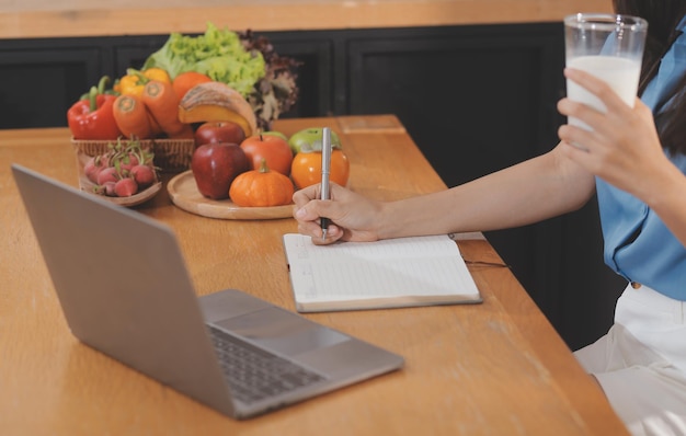 Una giovane donna con un bel viso in una camicia blu con i capelli lunghi che mangia frutta seduta in cucina a casa con un laptop e un taccuino per il relax Concept Vacation