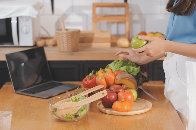 Una giovane donna con un bel viso in una camicia blu con i capelli lunghi che mangia frutta seduta in cucina a casa con un laptop e un taccuino per il relax Concept Vacation