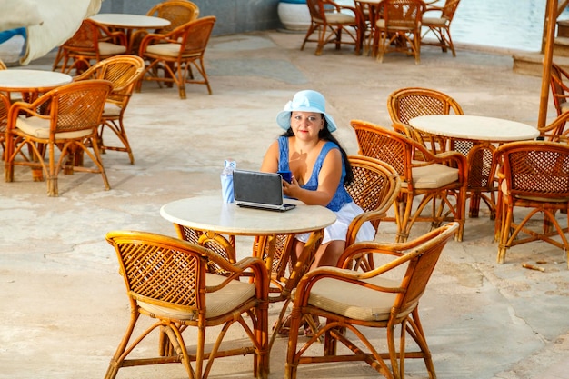 Una giovane donna con un cappello a un tavolo al mattino a bordo piscina beve un cocktail e lavora a distanza