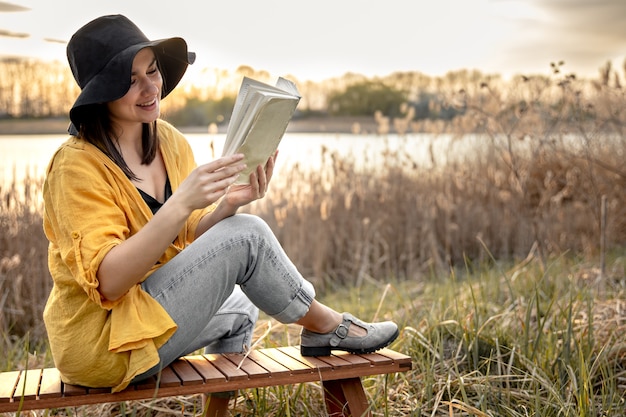 Una giovane donna con un cappello con un sorriso sul viso sta leggendo un libro seduta in riva al fiume al tramonto