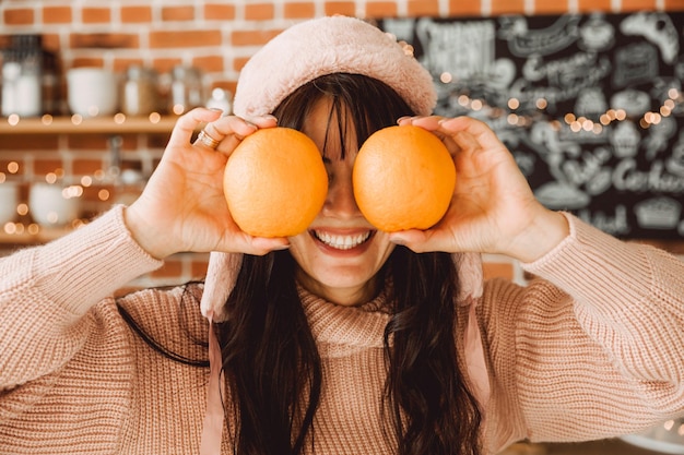 Una giovane donna con un cappello di pelliccia invernale tiene un'arancia vicino agli occhi. Cibo sano e concetto di stile di vita