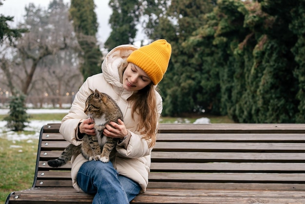 Una giovane donna con un cappello giallo siede su una panchina e tiene in grembo un gatto maculato in inverno Il concetto di buon trattamento degli animali e aiuto Posto per il tuo design