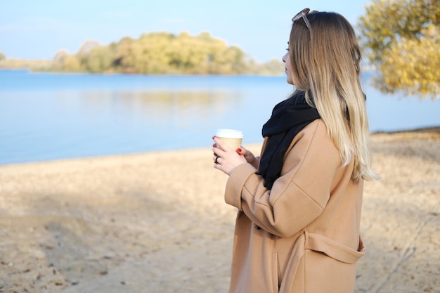 Una giovane donna con un cappotto marrone e una sciarpa nera sta con una tazza di caffè in mano e si gode il cielo azzurro e la vista sul lago Il concetto di armonia ed equilibrio