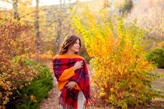 Una giovane donna con un maglione arancione si avvolge in una sciarpa in un parco autunnale