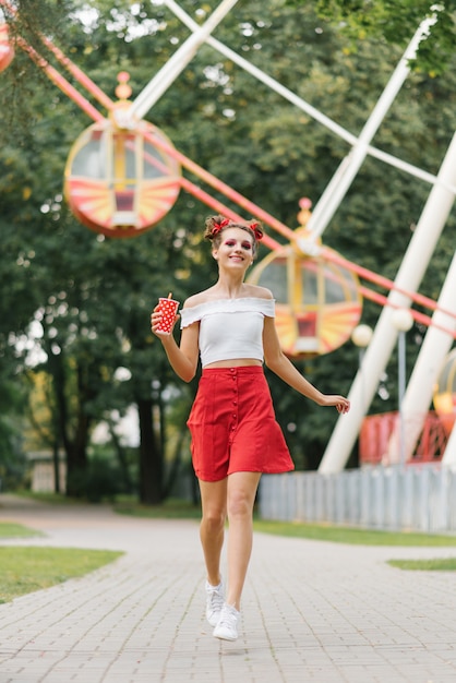 Una giovane donna con un trucco luminoso tiene in mano una tazza di carta rossa e attraversa un parco di divertimenti. Lei è sorridente e felice.