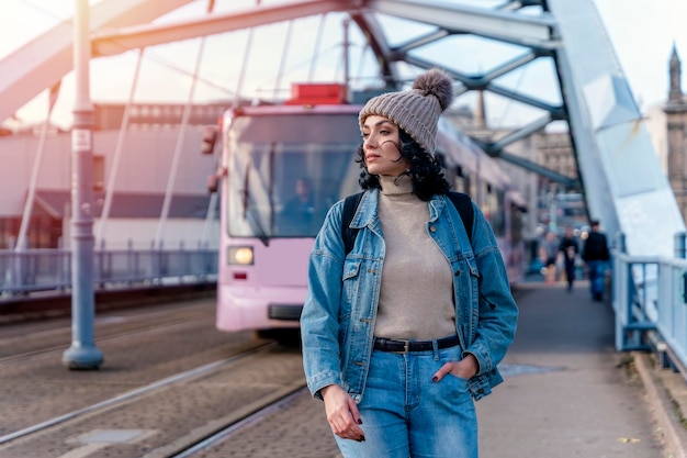 Una giovane donna con una giacca di jeans sta parlando al telefono e sta aspettando un tram alla fermata