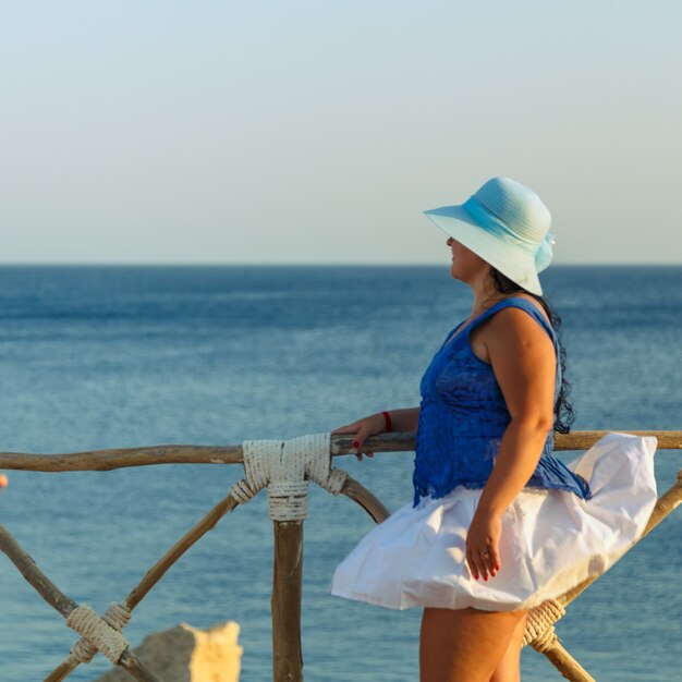 Una giovane donna con una gonna bianca e un cappello da sole si trova sulla riva sullo sfondo del mare al tramonto, guarda il mare. Foto quadrata