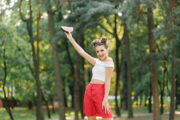 Una giovane donna con una gonna rossa e un top bianco si rallegra in estate e tiene in mano una fetta di anguria e sorride
