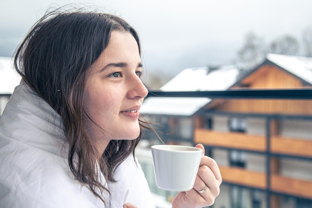 Una giovane donna con una tazza di caffè sul balcone in inverno in montagna