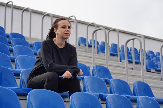 Una giovane donna con una tuta nera con i capelli lunghi si siede sullo stadio da sola e guarda una partita di sport
