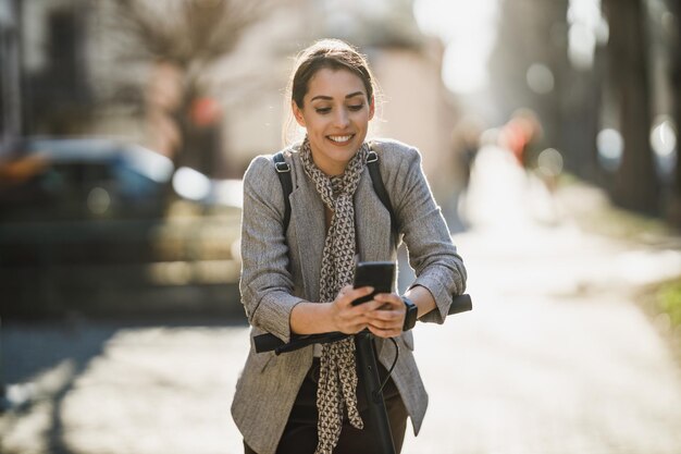 Una giovane donna d'affari che usa lo smartphone mentre va al lavoro con uno scooter elettrico.