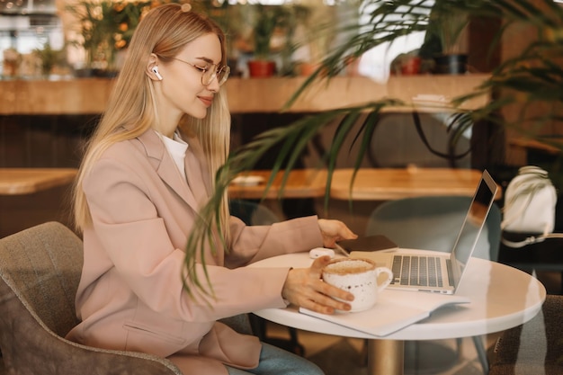 Una giovane donna d'affari con gli occhiali parla al telefono e lavora online utilizzando un laptop in un bar