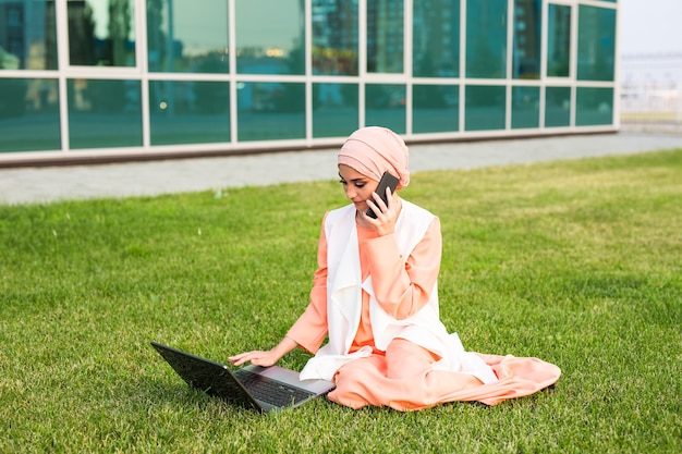Una giovane donna d'affari piuttosto musulmana in un parco mentre parla su smartphone guardando il laptop