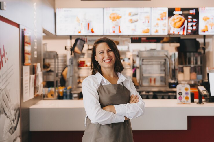 Foto una giovane donna d'affari sorridente in un grembiule si trova in un caffè di fronte a una vetrina, le braccia incrociate, guardando la telecamera.