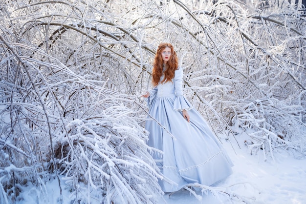 Una giovane donna dai capelli rossi, una principessa, cammina in una foresta invernale con un vestito blu. Gelo e neve sugli alberi.