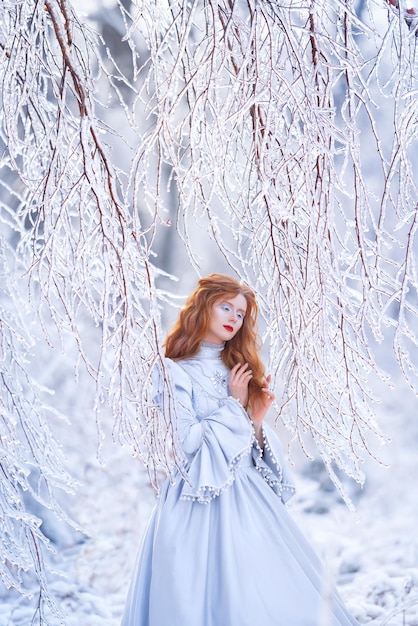 Una giovane donna dai capelli rossi, una principessa, cammina in una foresta invernale con un vestito blu. Gelo e neve sugli alberi.