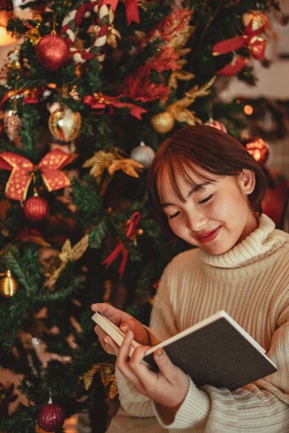 Una giovane donna davanti all'albero di Natale che legge un libro a casa con decorazioni natalizie.