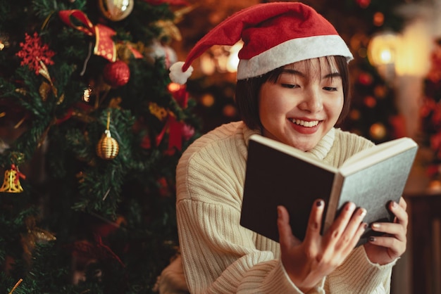 Una giovane donna davanti all'albero di Natale che legge un libro a casa con decorazioni natalizie.