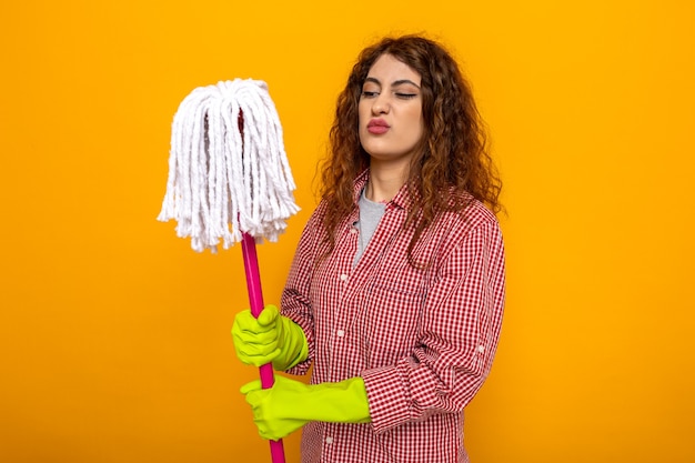 Una giovane donna delle pulizie scontenta che indossa guanti che tiene e guarda il mop