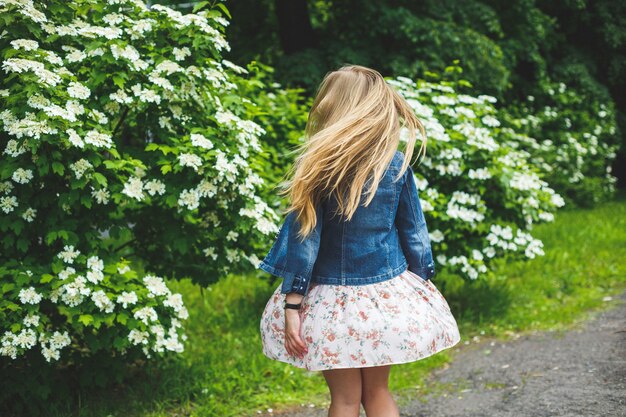 Una giovane donna di aspetto europeo con lunghi capelli biondi, vestita con un abito corto, si erge sullo sfondo di cespugli fioriti bianchi. Giornata di sole primaverile. Bellezza femminile naturale