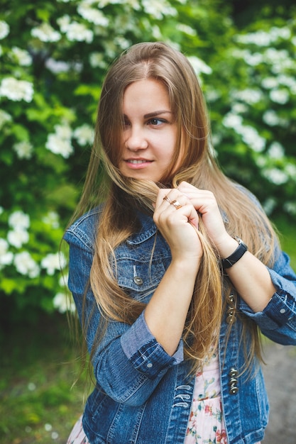 Una giovane donna di aspetto europeo con lunghi capelli biondi, vestita con un abito corto, si erge sullo sfondo di cespugli fioriti bianchi. Giornata di sole primaverile. Bellezza femminile naturale