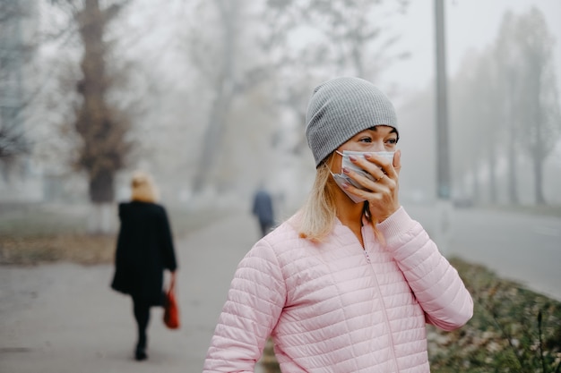 Una giovane donna è in piedi vicino alla strada con una maschera medica in autunno.