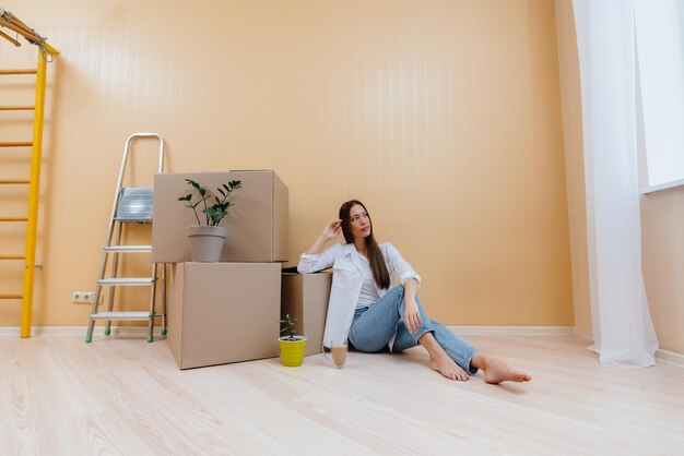 Una giovane donna è seduta per terra davanti agli scatoloni e beve caffè, gioendo e godendosi il nuovo appartamento dopo il trasloco. Inaugurazione della casa, consegna e trasporto merci.