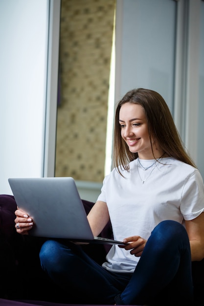 Una giovane donna è seduta sul divano e lavora su un laptop in remoto dal lavoro. Ragazza con un computer in ginocchio che guarda il monitor