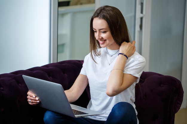 Una giovane donna è seduta sul divano e lavora su un laptop in remoto dal lavoro. Ragazza con un computer in ginocchio che guarda il monitor