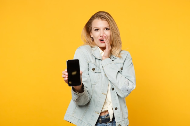 Una giovane donna eccitata in abiti di jeans racconta qualcosa con un gesto della mano, tenendo il telefono cellulare con uno schermo vuoto vuoto isolato su sfondo giallo arancione. Concetto di stile di vita delle persone. Mock up spazio di copia.