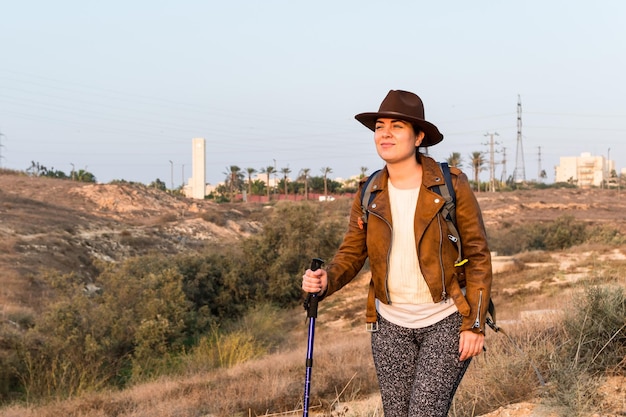 Una giovane donna escursionista con una giacca di pelle, un cappello, uno zaino e un bastone di tracciamento cammina attraverso un campo
