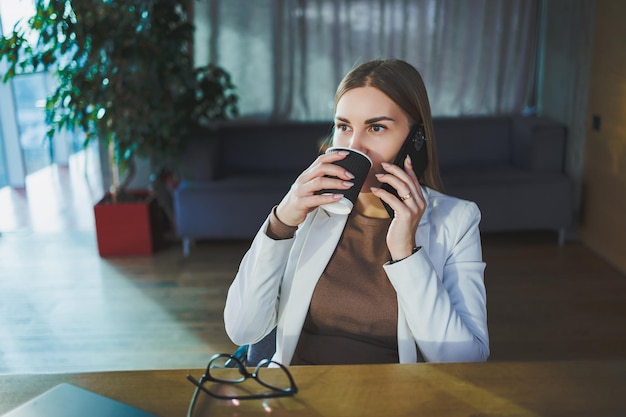 Una giovane donna felice con lunghi capelli biondi in abiti eleganti è seduta a un tavolo con un laptop che si gode una tazza di caffè fresco e parla al telefono Lavora in un ufficio moderno con grandi finestre