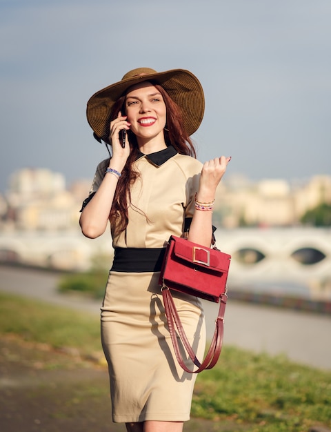 Una giovane donna felice con un cappello a tesa larga cammina nel parco in estate con una borsa e parla al telefono