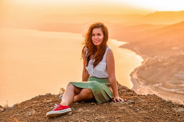 Una giovane donna felice con una gonna è seduta sulla cima di una scogliera di montagna sotto il cielo del tramonto