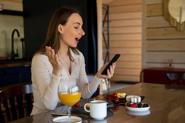 Una giovane donna felice è seduta in cucina a casa e usando un telefono cellulare ha ricevuto un messaggio buone notizie