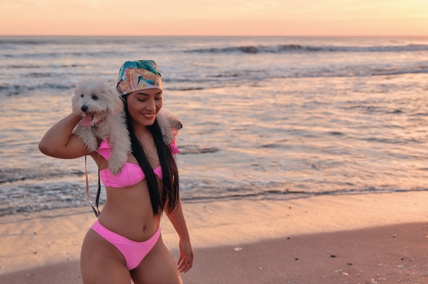 Una giovane donna felice in bikini che porta il suo simpatico cucciolo peloso sulle spalle in riva al mare
